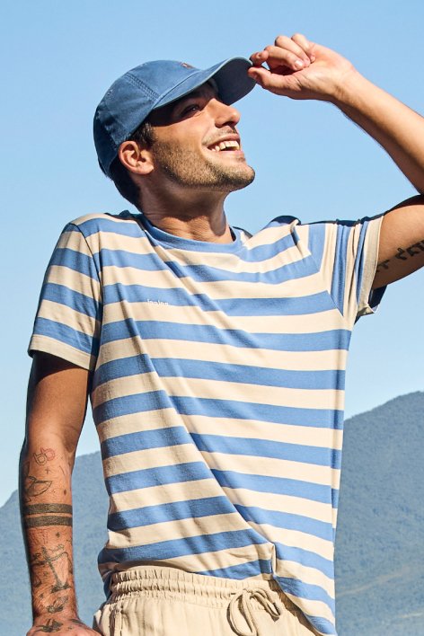 Homem usando uma camisa listrada azul e branca, acompanhado de um chapéu, em um ambiente ao ar livre.