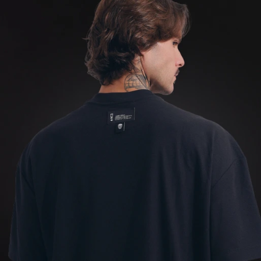Detalhe das costas de um modelo masculino vestindo uma camiseta preta lisa da marca MCD. Destaque para uma pequena etiqueta preta costurada logo abaixo da gola, contendo o logotipo da marca e um pequeno ícone de caveira.