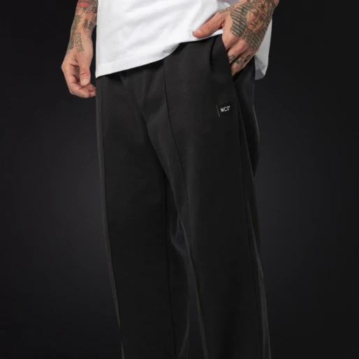 Detalhe de um modelo vestindo uma calça preta de modelagem reta e solta da marca MCD. O modelo está com a mão no bolso, revelando uma pequena etiqueta preta com a inscrição 